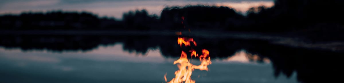 The top of a bonfire flame, against a background of trees reflected in a lake.